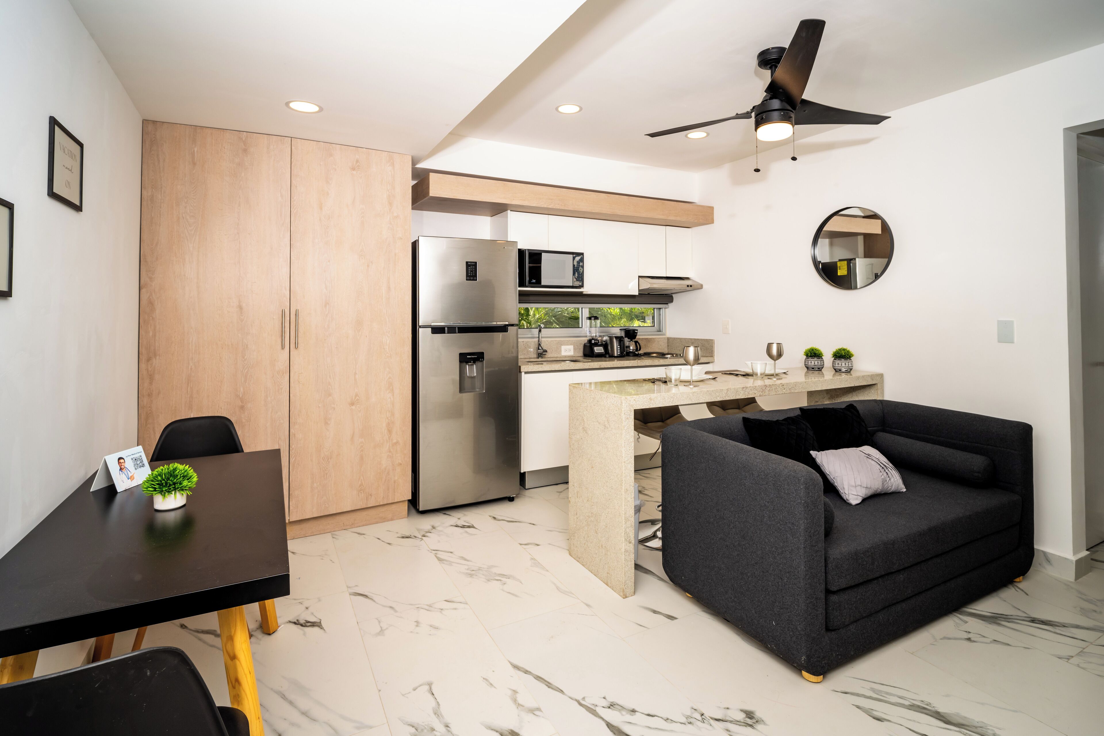 A modern kitchen with a refrigerator, microwave, and coffee maker. There is a dining table with chairs and a sofa.