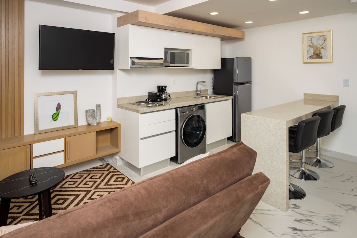 A compact kitchen with a microwave, sink, and refrigerator. There is a sofa and a small table with a remote control.