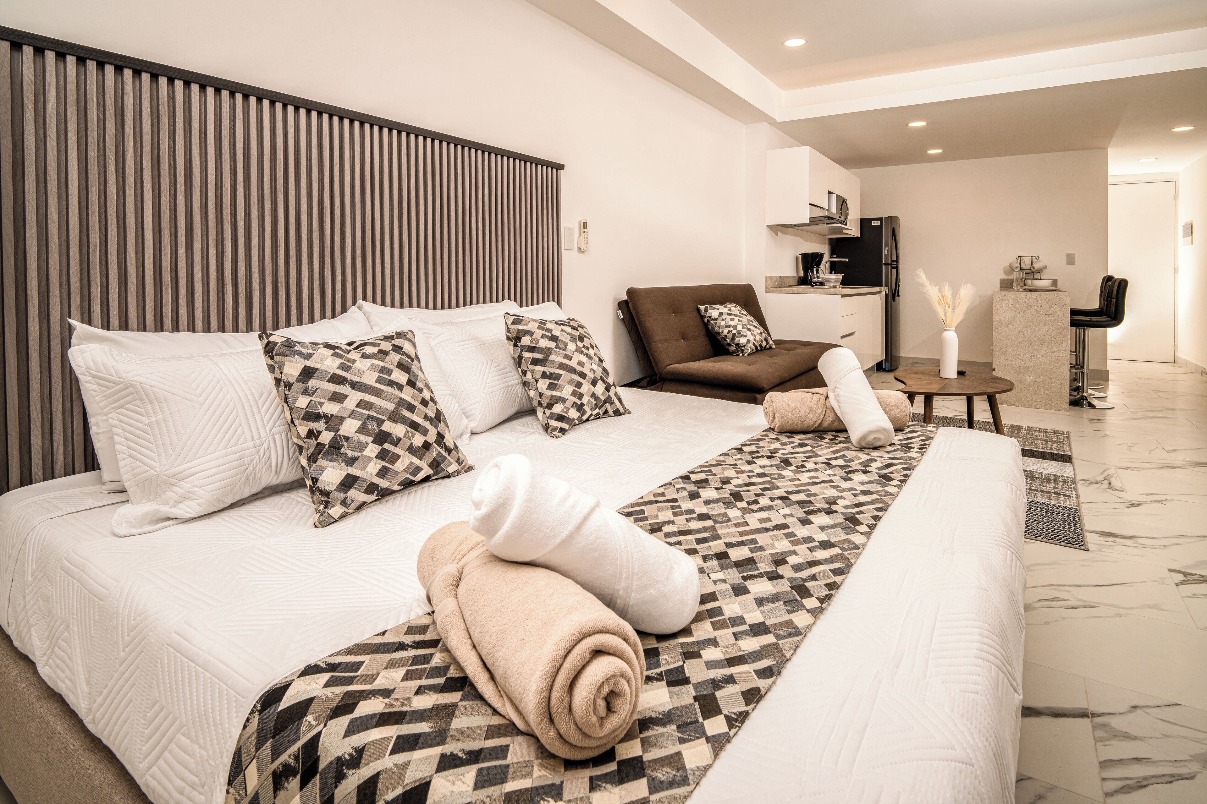 Une chambre d’hôtel moderne avec un grand lit, un canapé, une petite table et un coin cuisine à l’arrière-plan.