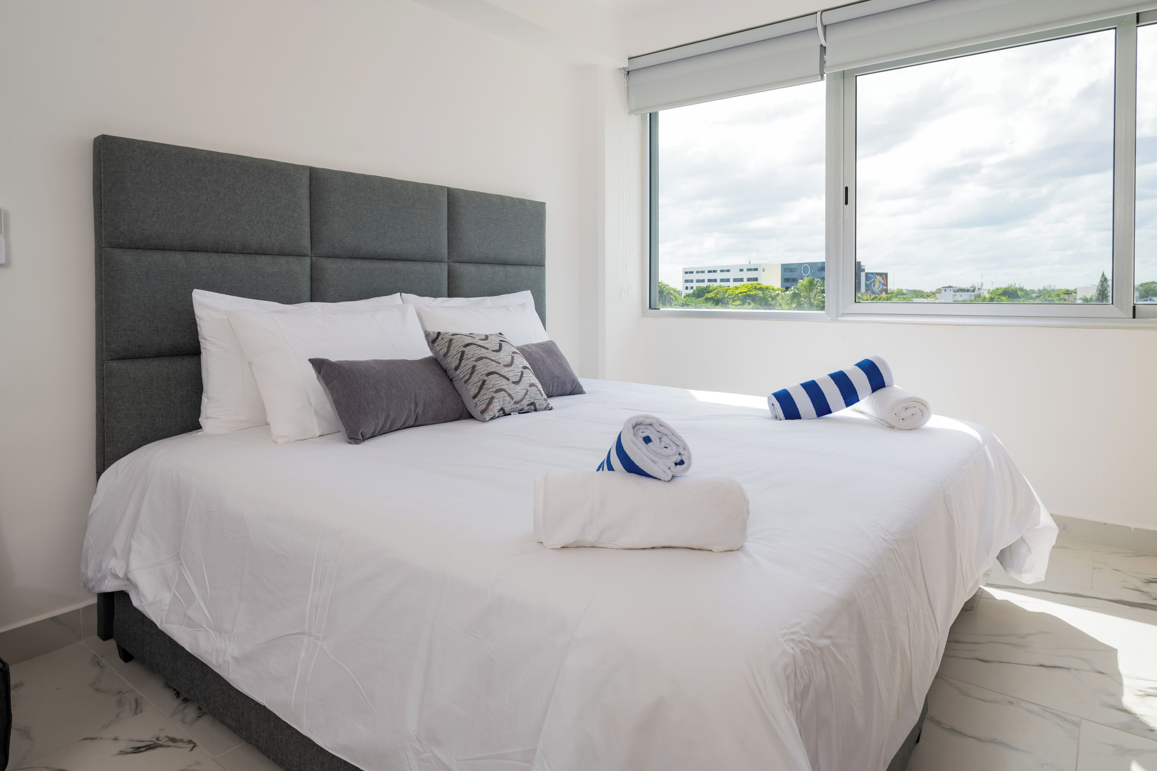 A neatly made bed with a grey headboard, white bedding, and a rolled towel.