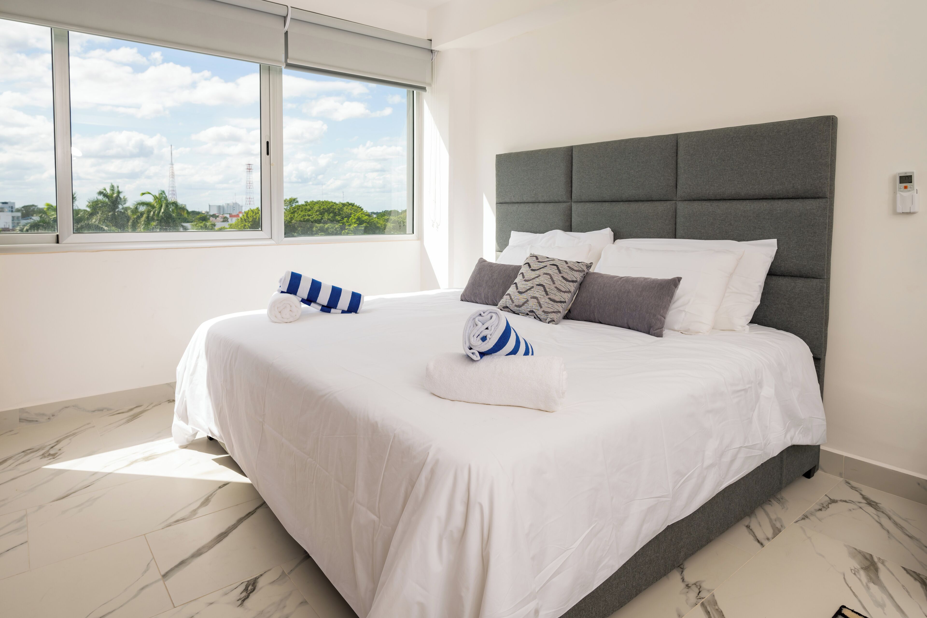 Een moderne hotelkamer met een groot bed, een grijs gestoffeerd hoofdeinde, witte beddengoed en een uitzicht op de skyline van de stad door grote ramen.