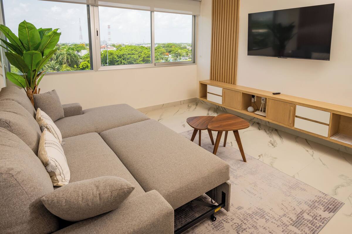 A modern living room with a sectional sofa, a flat-screen TV, a wooden console table, and a large window offering a city view.