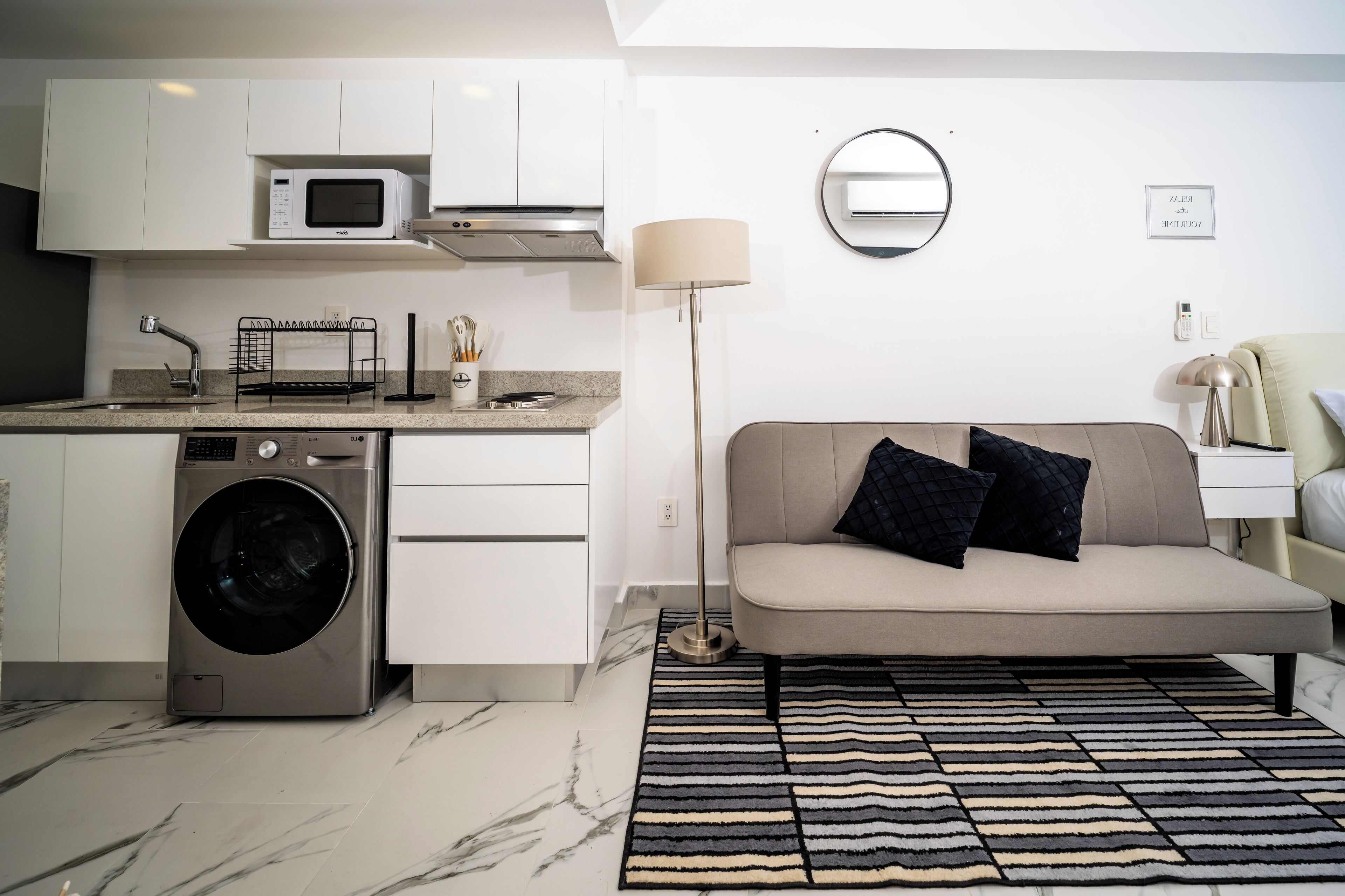 A compact kitchen with a washing machine, a microwave, and a sink. There's a sofa with cushions and a floor lamp.