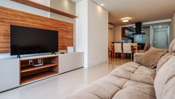 Ein modernes Wohnzimmer mit großem Flachbildfernseher, einer Holz-Unterhaltungseinheit, einem beigefarbenen Sofa und einer Küchenzeile im Hintergrund.