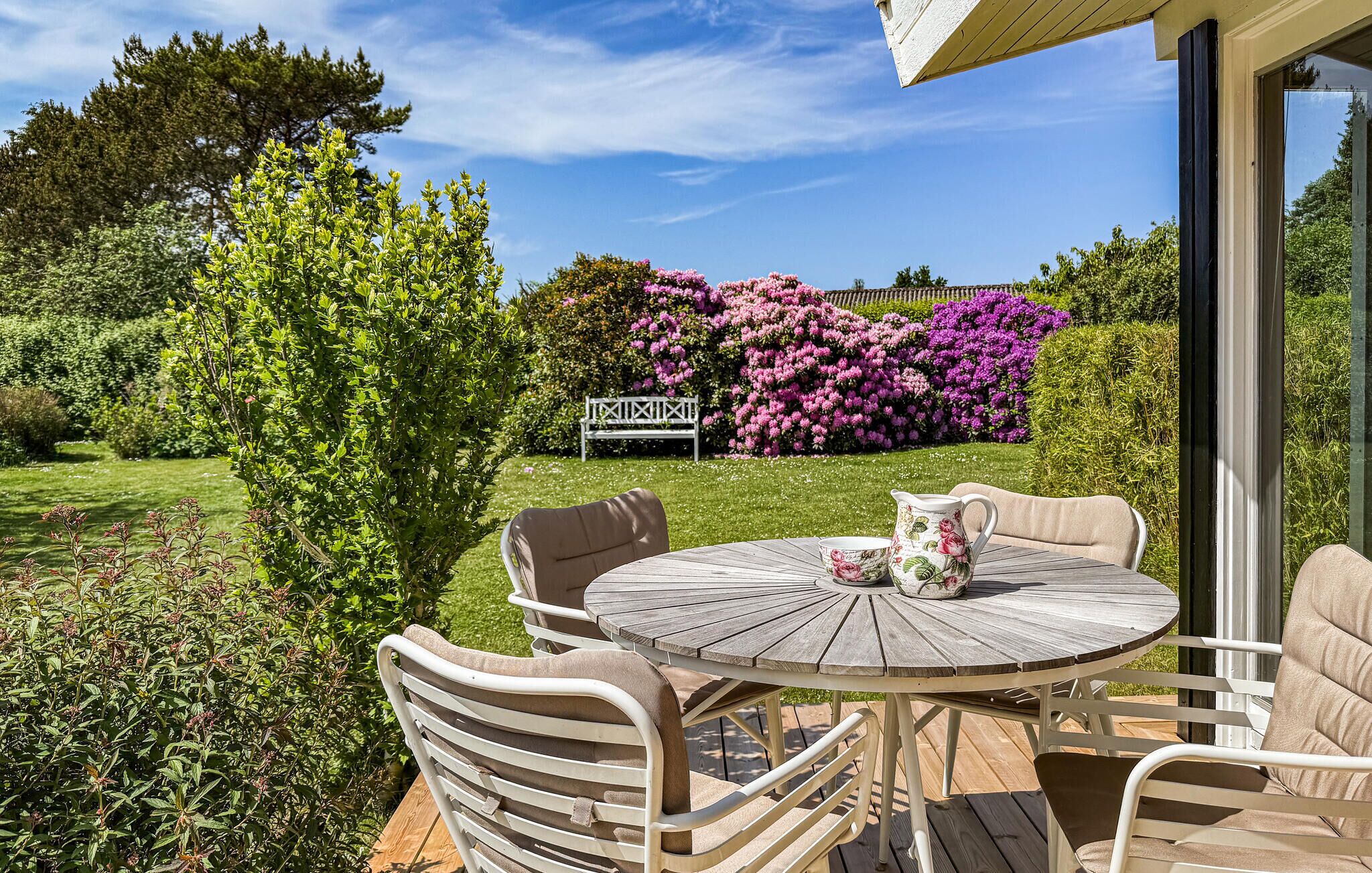 An outdoor patio with a round wooden table and white cushioned chairs, surrounded by lush greenery and flowering bushes.