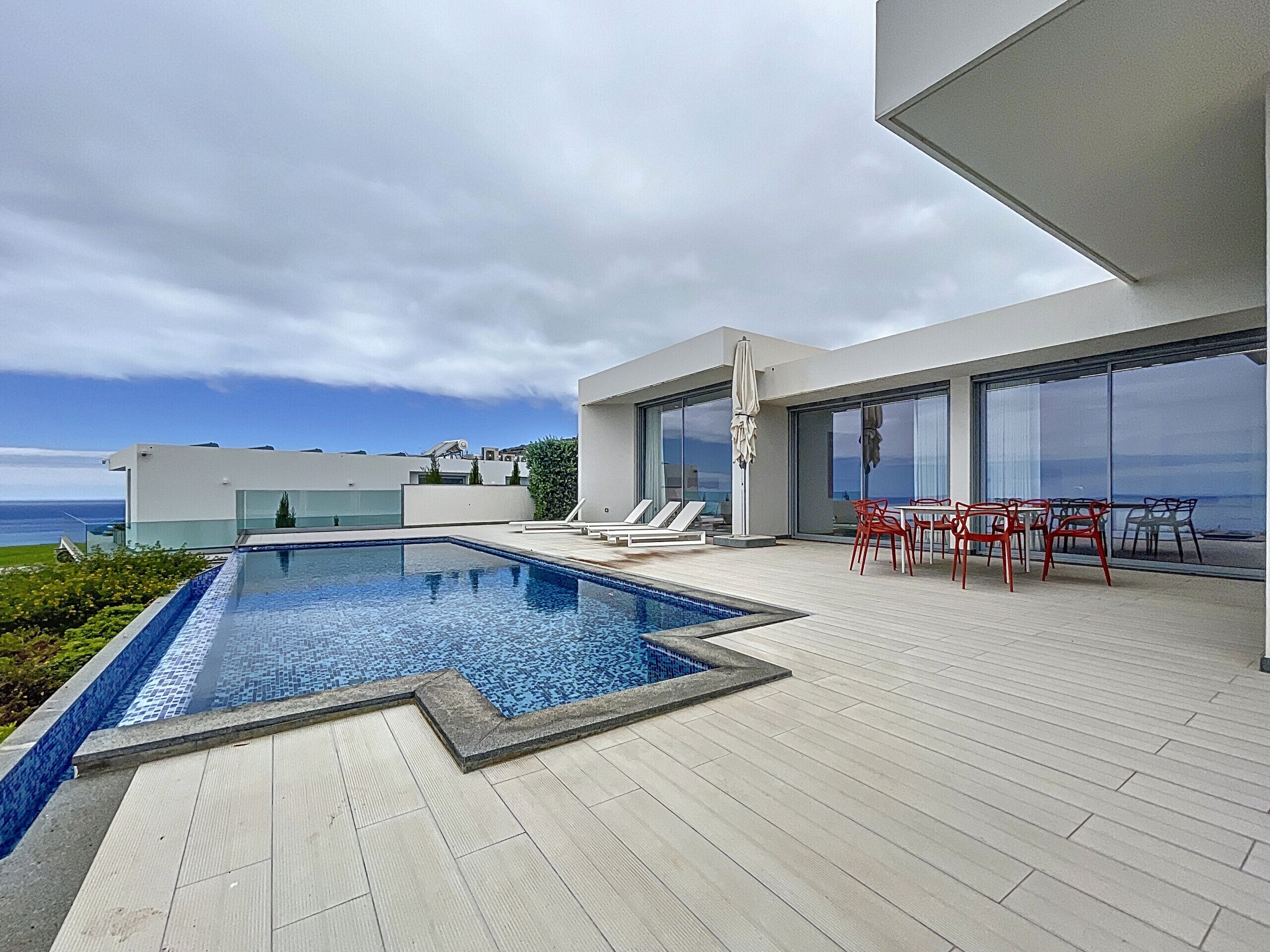 A modern outdoor pool area with a view of the sea, a dining area with red chairs, and a large glass door leading to a covered patio.