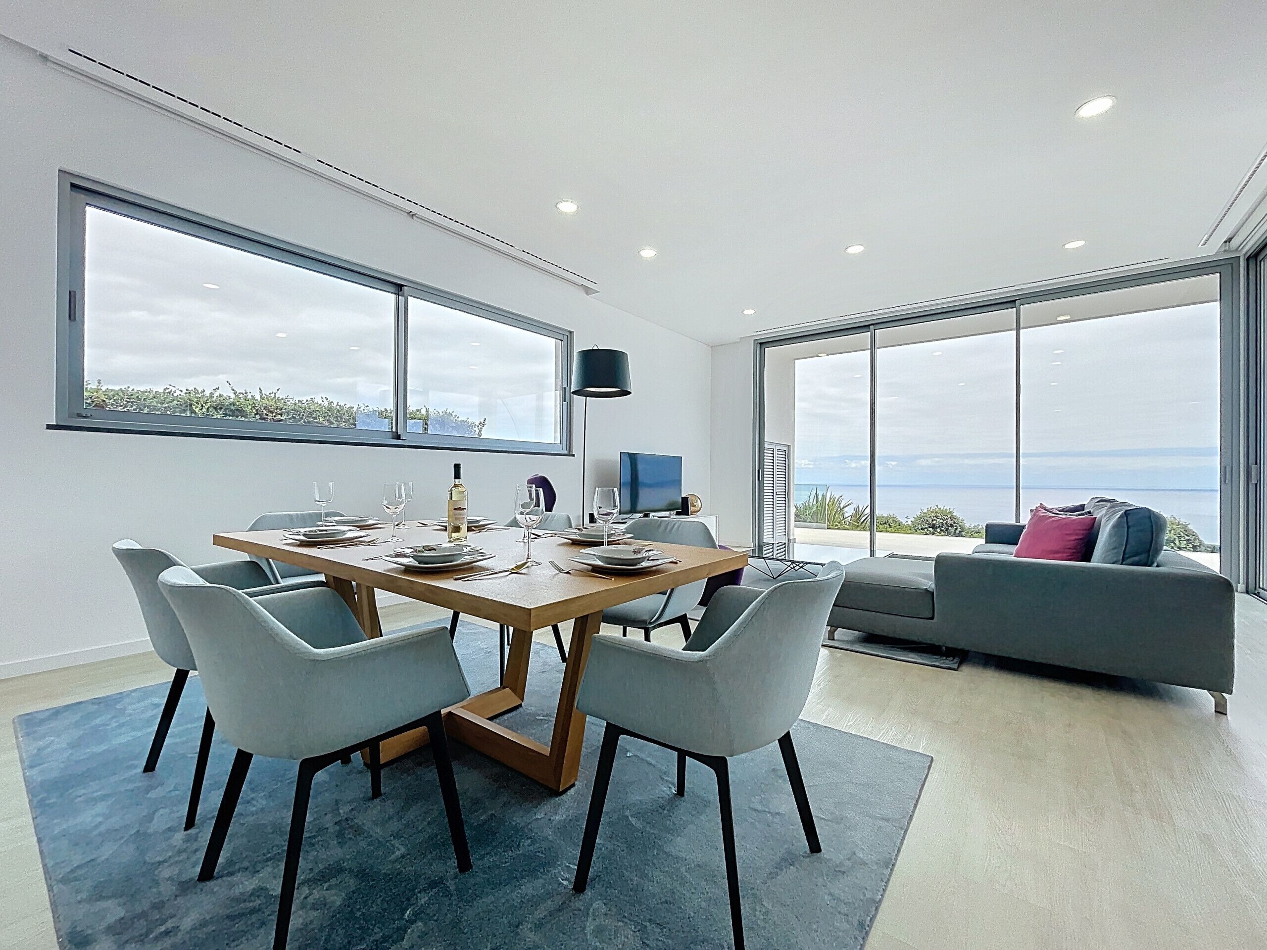 Un espace repas moderne avec une table en bois, des chaises grises et une grande fenêtre donnant sur la mer.