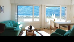 A living room with a large window overlooking a mountainous landscape. There is a teal sofa, a small round table, and a dining table with chairs. - Stunning Apartment with 180 Degree Panoramic Views to Lake Maggiore (Bellinzona)