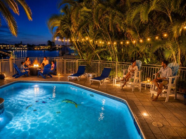 A swimming pool with lounge chairs and a fire pit area, surrounded by palm trees and string lights.