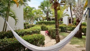 A hammock in a lush garden with palm trees and a pathway leading to a building.