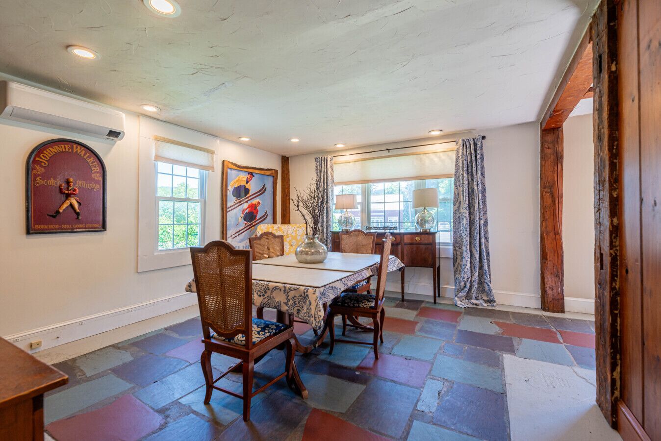 Comedor con mesa de madera, sillas de mimbre y mantel estampado. Hay dos ventanas con cortinas, un cuadro en la pared y un logotipo en la pared.