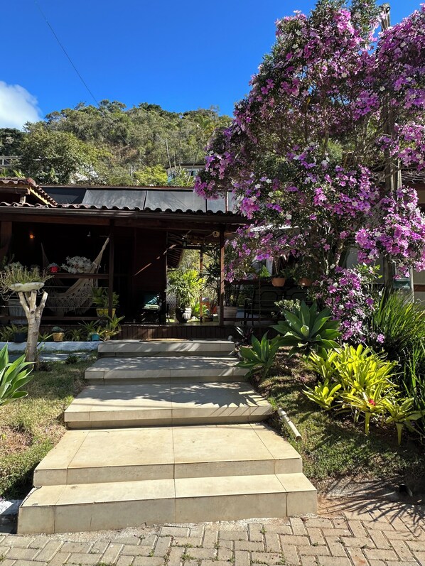 A house with a porch, steps leading to the entrance, and a garden with various plants and a flowering tree. - Pousada Dulce Ville (Domingos Martins)