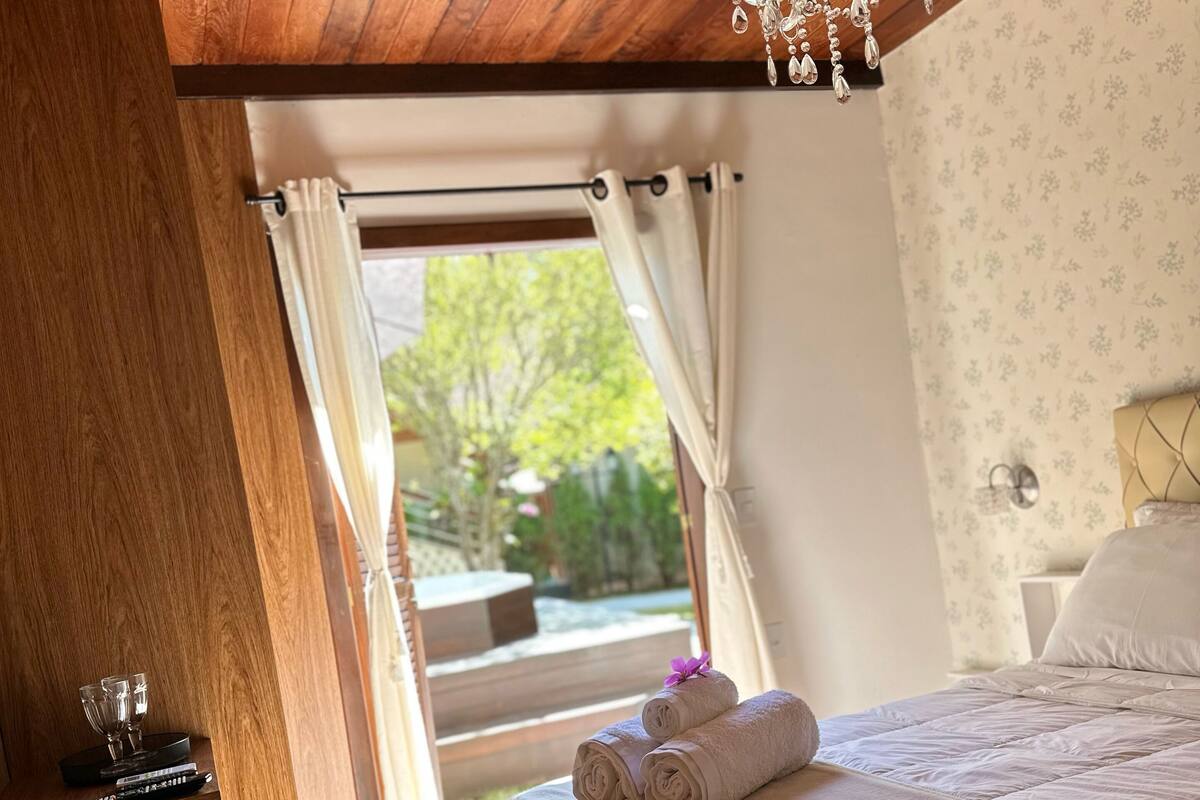 A bedroom with a wooden ceiling, a chandelier, a bed with a white duvet, and a view of greenery outside.