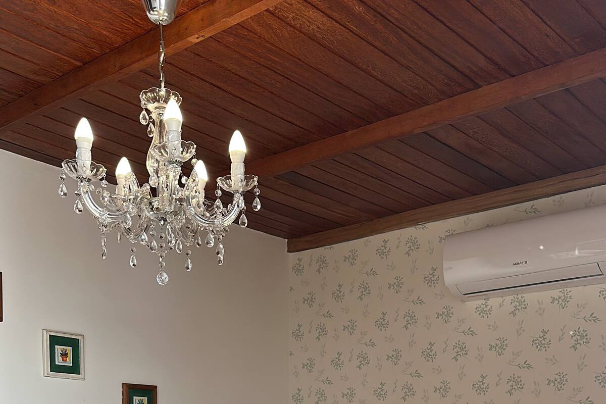A bedroom with a wooden ceiling, a chandelier, a bed with a beige headboard, and an air conditioning unit on the wall.