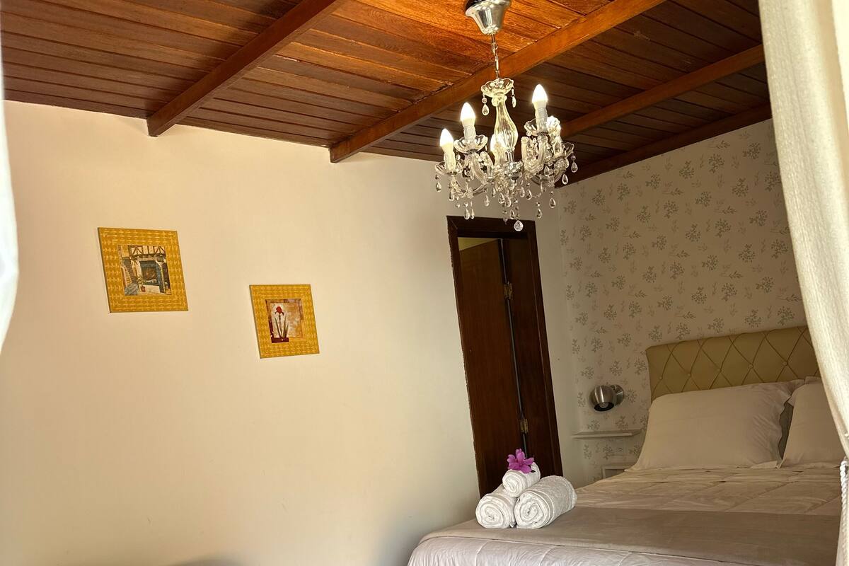 A bedroom with a bed, a red chair, a chandelier, and two framed pictures on the wall.