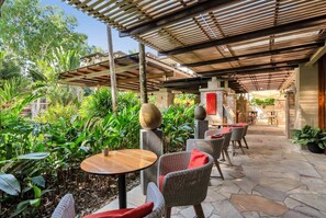 Un espace extérieur aménagé en terrasse, avec des tables rondes et des chaises en osier, entouré d’une végétation luxuriante et agrémenté d’une pergola en bois.