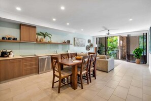 A modern kitchen with wooden cabinets, a dining area with a wooden table and chairs, and a sliding glass door leading to a patio with greenery.