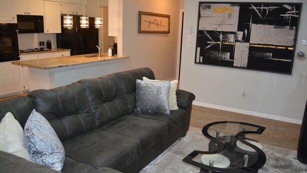 A modern living room with a dark leather sofa, a glass coffee table, and a kitchen area in the background. - Fenced Backyard + Parking - Near Six Flags + AT&T (Forest Hill)