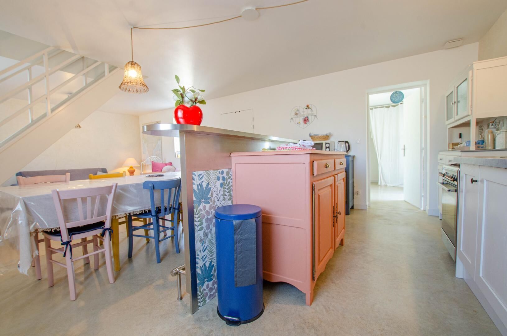 Une cuisine chaleureuse avec un coin repas, comprenant un escalier, une poubelle et un vase en forme de cœur posé sur le comptoir.