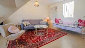 Een gezellige woonkamer met een bank, een fauteuil, een klein rond tafeltje en een bed met een rood geweven vloerkleed.