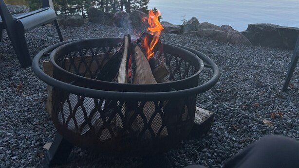 Ein Lagerfeuer mit Holzscheiten, umgeben von Kies, in der Nähe eines ruhigen Sees mit Bäumen und einem Stuhl im Vordergrund.