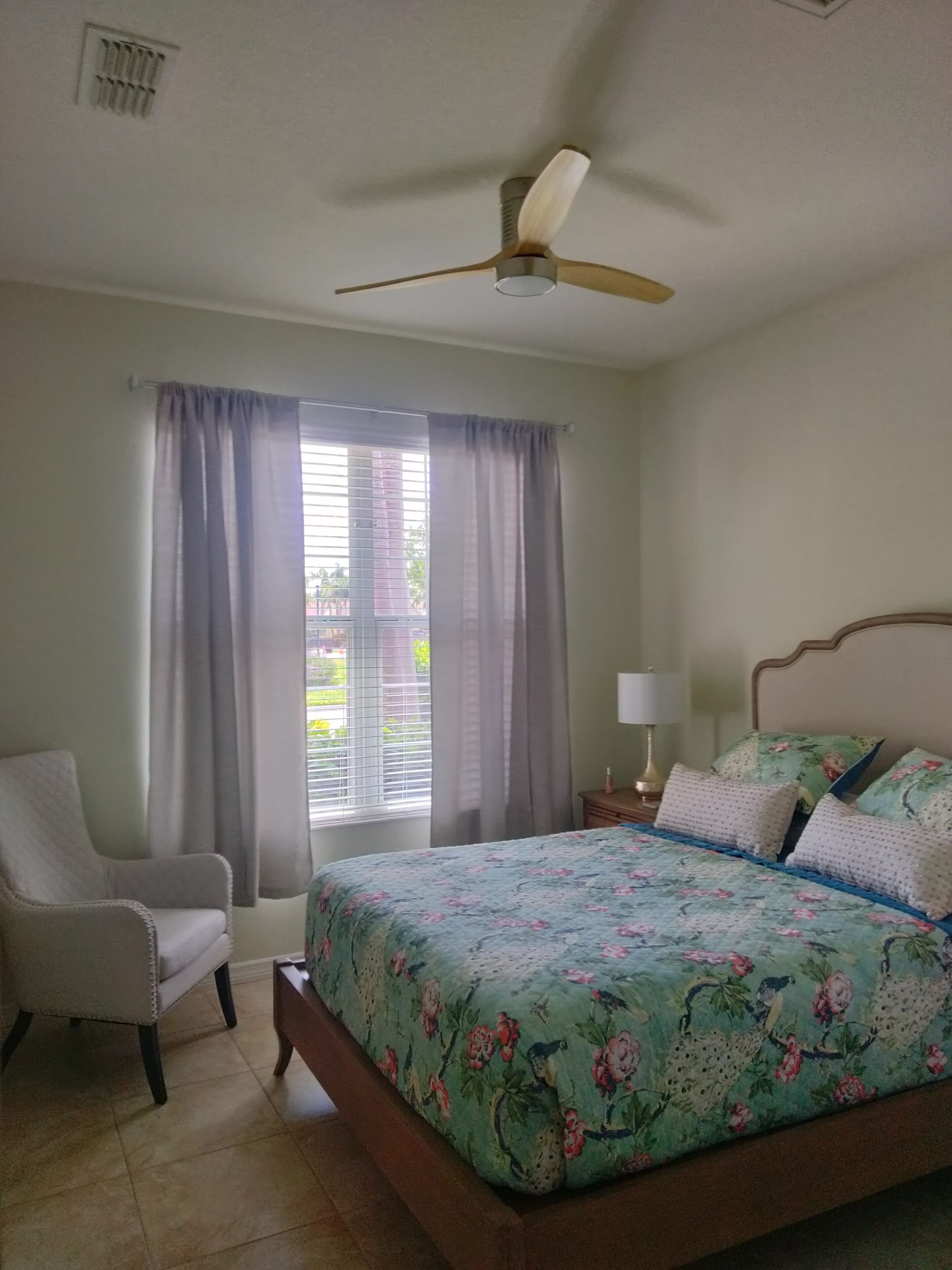 A bedroom with a bed, a chair, a nightstand with a lamp, and a ceiling fan.