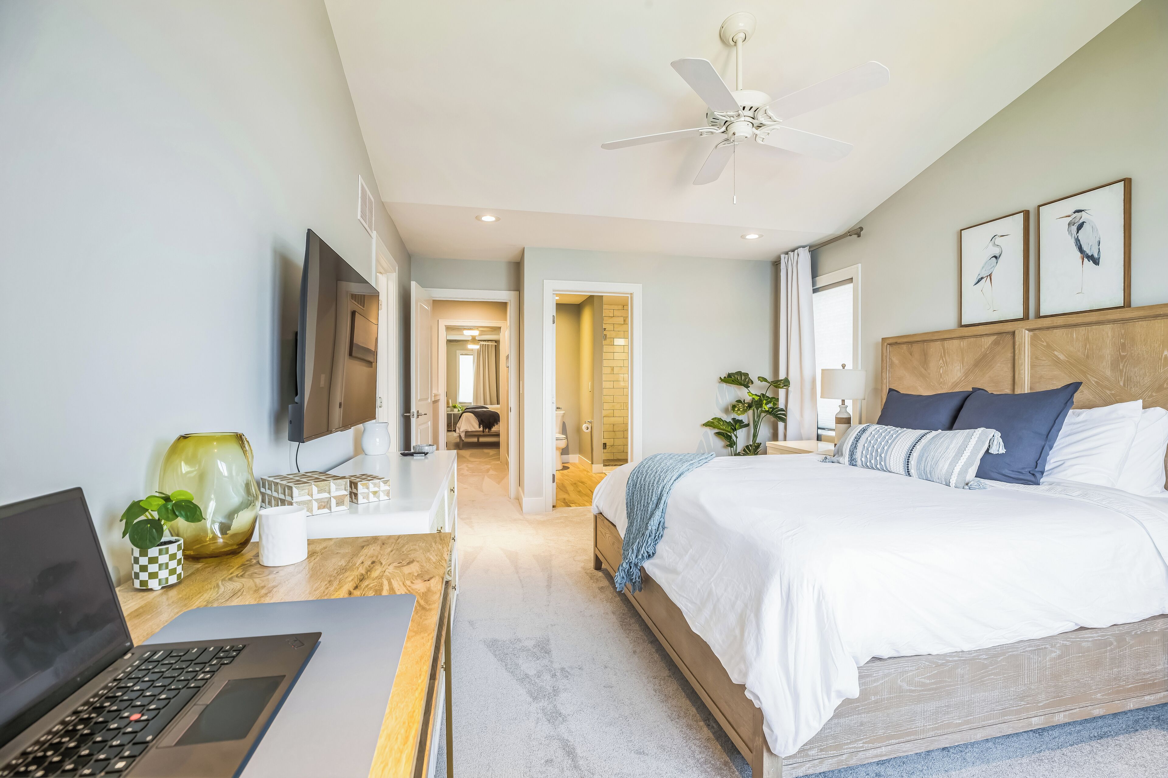 Ein modernes Schlafzimmer mit Bett, Schreibtisch mit Laptop, Fernseher und Deckenventilator.