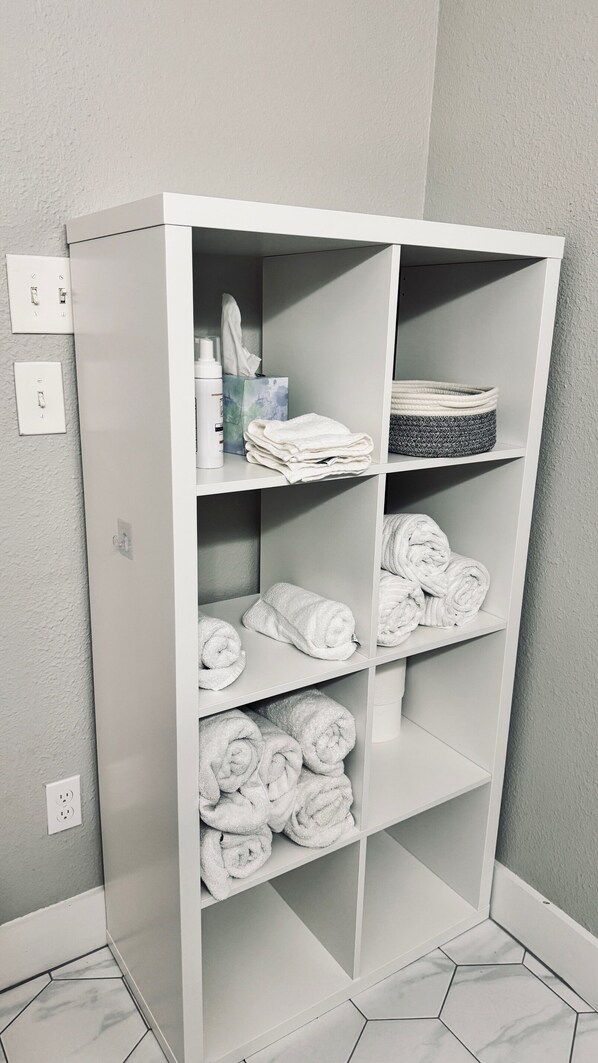 A white storage unit with multiple shelves, some holding folded towels and a basket.