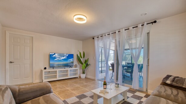 A living room with a television, sofa, coffee table, and a view through large windows. - VILLA BEACH PARADISE – HEATED POOL & SPA | WATER VIEW (Bonita Springs)
