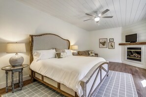 Una camera da letto con un letto di legno, due comodini con lampade, una ventola a soffitto, un camino e stampe botaniche incorniciate alle pareti.