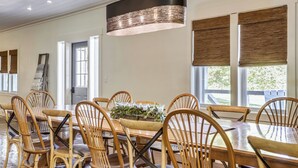 Un comedor con una mesa y sillas de madera, una lámpara colgante y ventanas amplias con persianas.