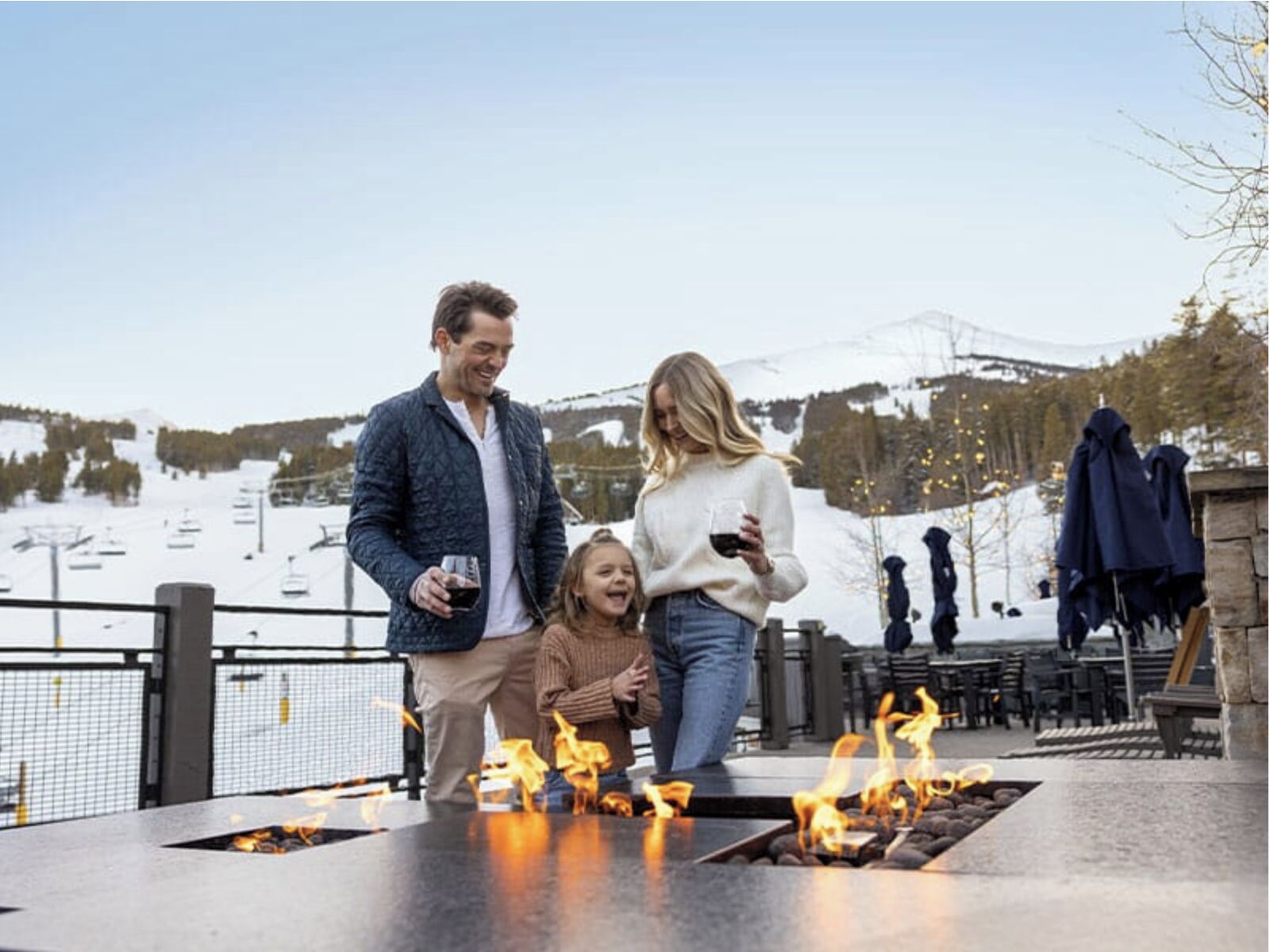 A family enjoying a fire feature outdoors with a ski slope in the background.