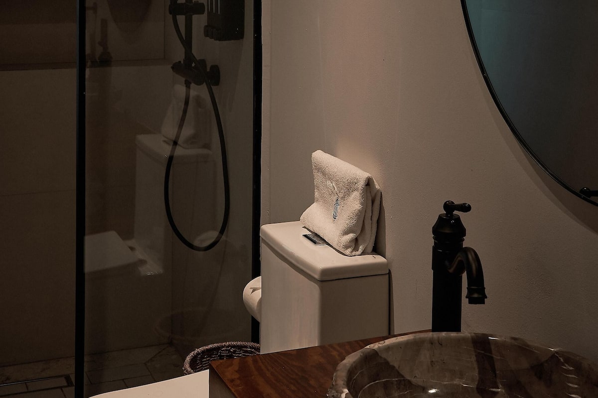 Une salle de bain dotée d’une douche à parois de verre, de toilettes blanches, d’un meuble-lavabo en bois avec un vasque en marbre et d’un grand miroir.