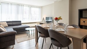 Un salon moderne avec un canapé, une table à manger pour quatre personnes et une grande fenêtre équipée de stores.