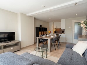 A modern living room with a flat-screen TV, grey sofas, a glass coffee table, and a dining area with a white table and chairs.