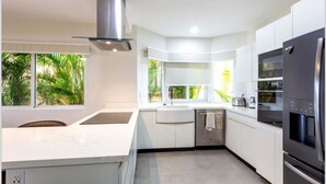 Una cuina moderna amb armaris blancs, electrodomèstics d'acer inoxidable i un gran finestral amb vista a vegetació.
