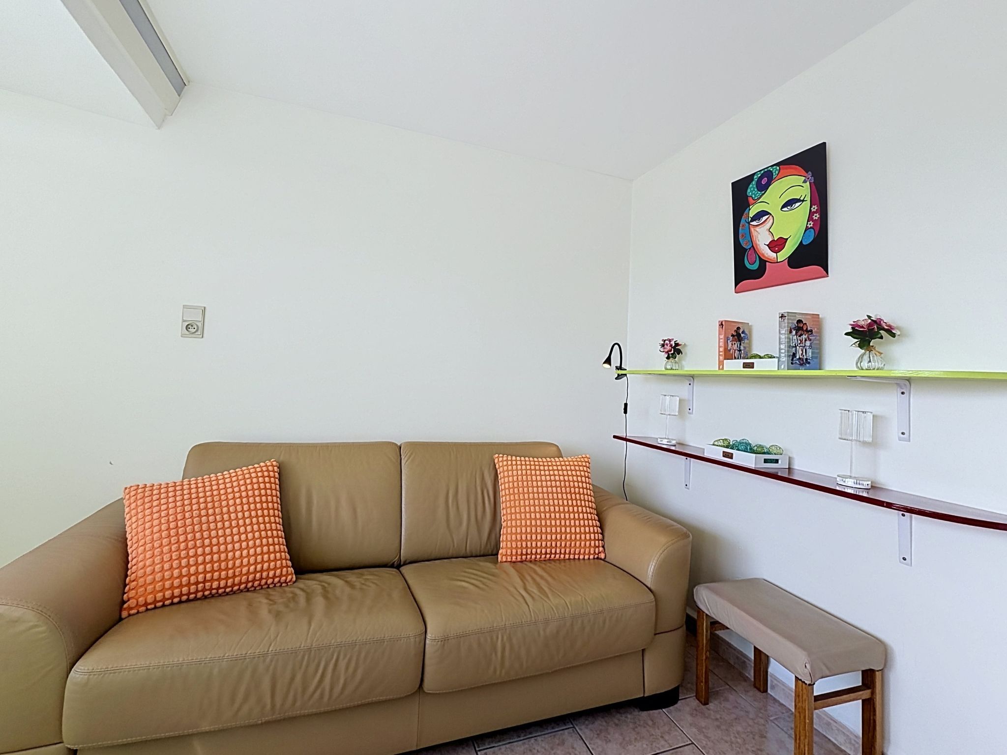Ein Wohnzimmer mit einem beigen Sofa, zwei orangefarbenen Kissen, einer Holzbank, einem an der Wand befestigten Regal mit Büchern und Blumen sowie einem farbenfrohen Gemälde.