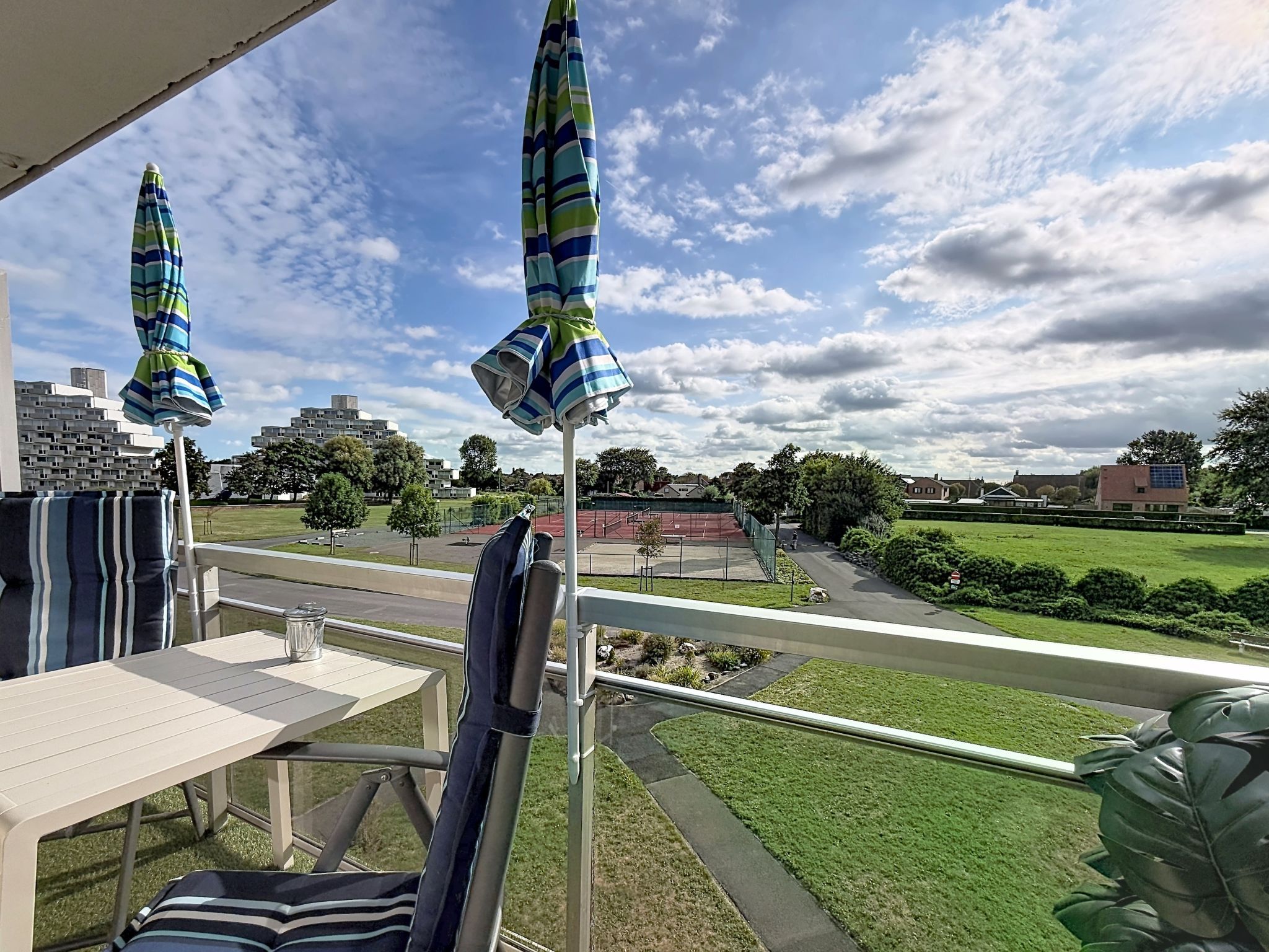 Un balcon avec une table et des chaises, deux parasols à rayures et une vue sur un parc verdoyant et des immeubles.