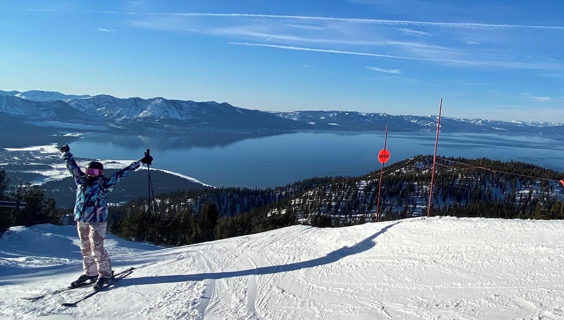 Een persoon skiënd op een besneeuwde helling, met een helder uitzicht op een meer en bergen op de achtergrond.