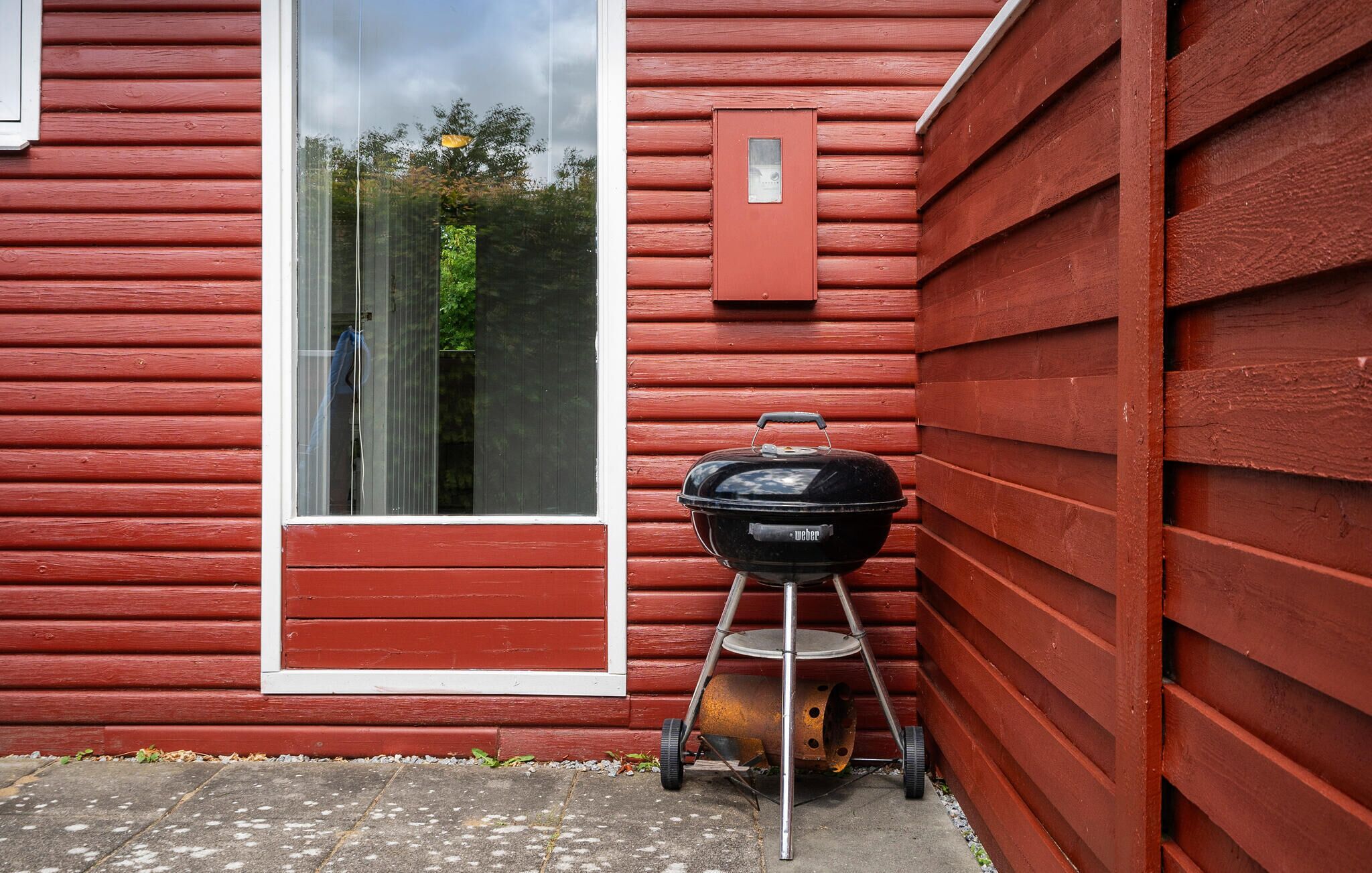 A red wooden house with a large window and a black Weber grill with a wheelbarrow in front.