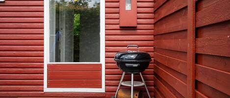 A red wooden house with a large window and a black Weber grill with a wheelbarrow in front.