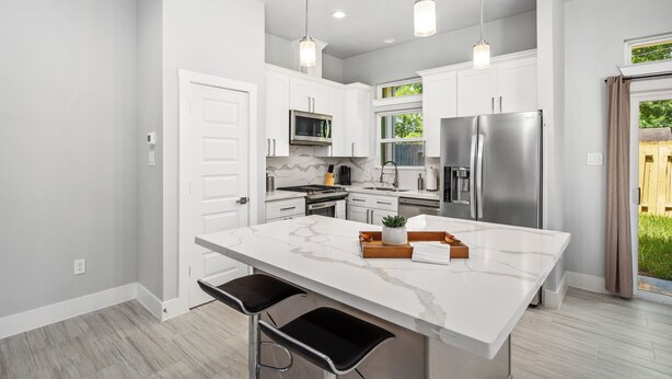 A modern kitchen with white cabinetry, a large island, stainless steel appliances, pendant lighting, and a view of greenery outside. - Elegant and beautiful 3-bedroom townhome near downtown Houston with WiFi, AC (Houston)