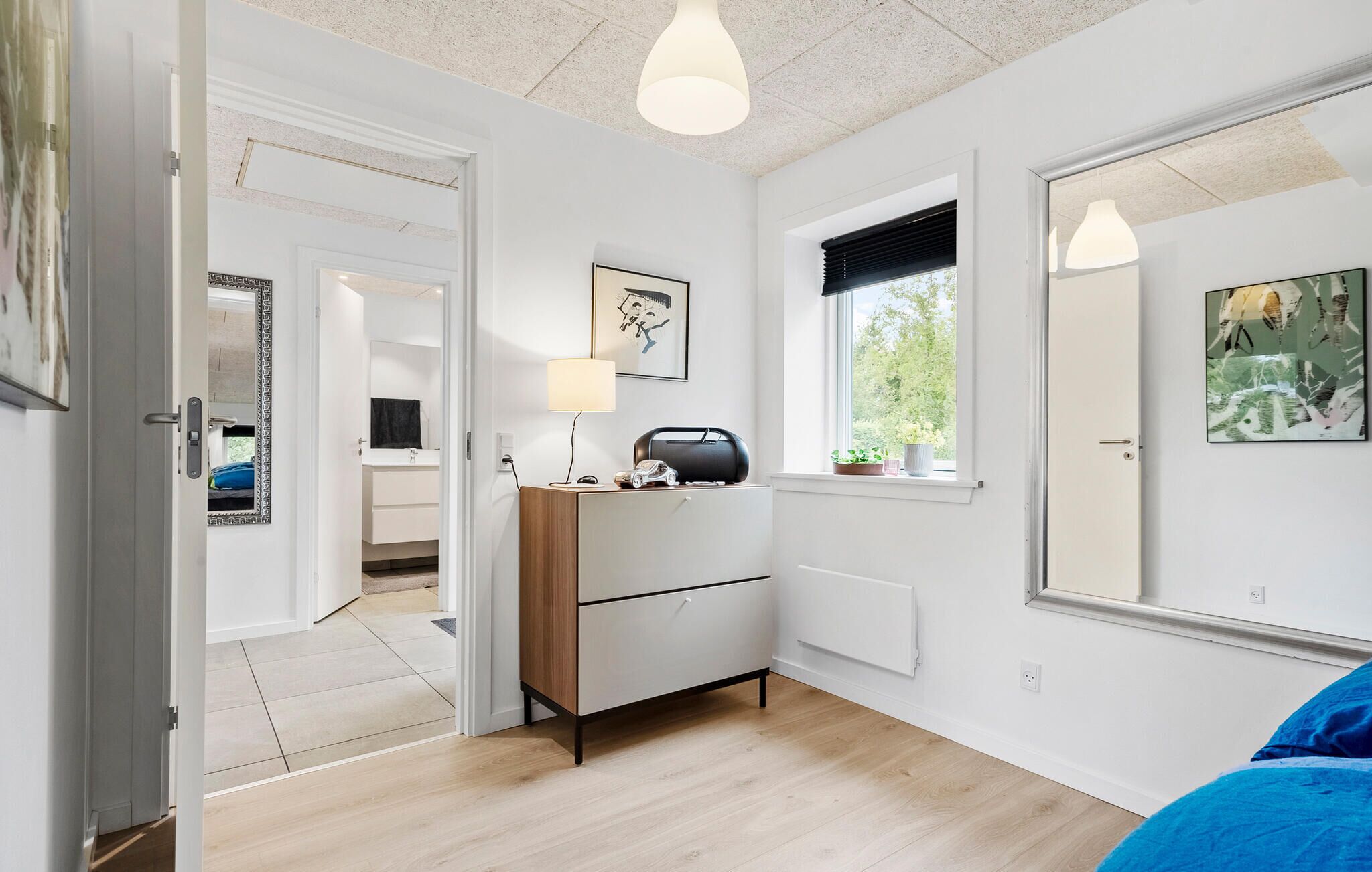 A modern bedroom with a wooden dresser, a bedside table with a lamp, a window with a view of greenery, and a door leading to another room.