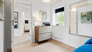 Une chambre moderne avec une commode en bois, une table de chevet avec une lampe, une fenêtre donnant sur un paysage verdoyant et une porte menant à une autre pièce.