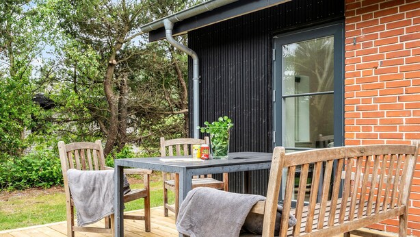 Une terrasse extérieure en bois avec une table et des chaises, un vase en verre contenant des plantes vertes, et un bâtiment en briques en arrière-plan.