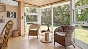 Quarto com piso de madeira, cadeiras de vime, uma mesa redonda e grandes janelas com vista para o exterior.