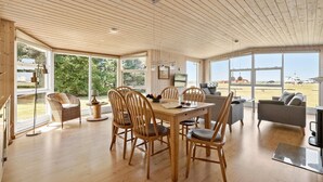 Quarto amplo com pisos de madeira e teto alto, com área de jantar incluindo cadeiras e mesa de madeira, um sofá, e janelas grandes que oferecem vista para o exterior.