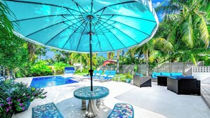 Een zwembadterras met een grote parasol, ligbedden en een tafel en bank met mozaïektegels.