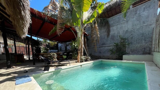 A swimming pool with clear water, surrounded by palm trees and a thatched roof structure. - Casa Firefly (Puerto Escondido)