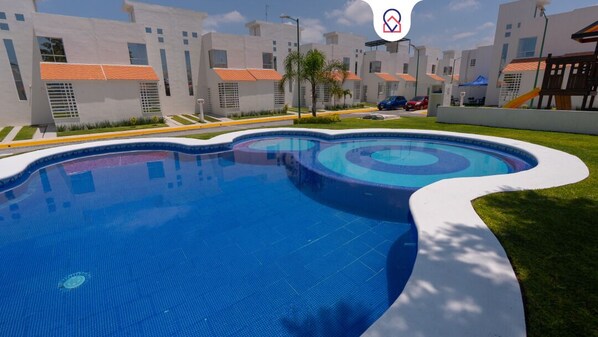 A swimming pool with a wavy edge, surrounded by a well-maintained lawn and a row of modern buildings with orange roofs. - FFamily House & Water Park Tlayecac 3 BR (Ayala)